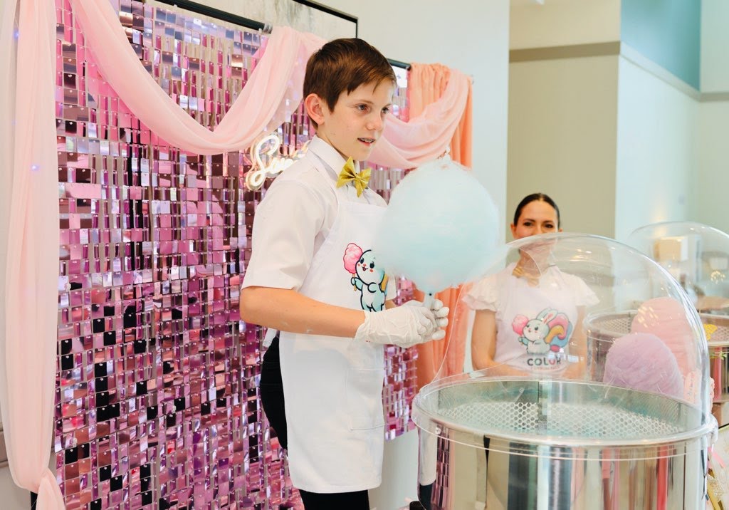 Liam holding blue cotton candy beside a cotton candy machine with a pink sequin backdrop